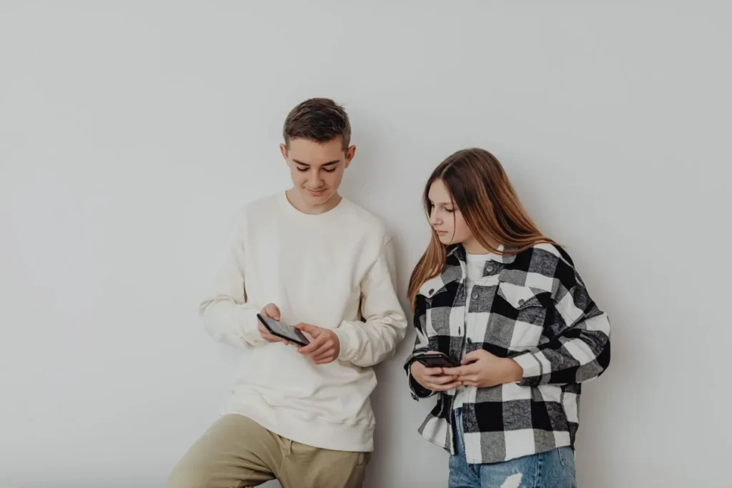 Teenage boy and girl standing against a wall, each using a smartphone
